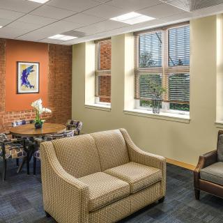 A cozy lounge area with a sofa, chairs, and a table adorned with flowers, featuring large windows and wall art.