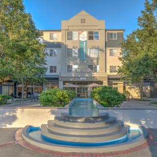 A modern building with a decorative fountain in front, surrounded by trees and greenery under a clear blue sky.
