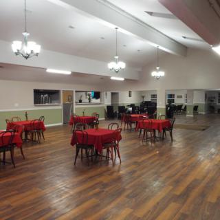 A spacious dining hall with tables covered in red tablecloths and wooden flooring.