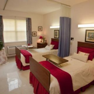 A hospital room with twin beds separated by a curtain, featuring red bedding and bright lighting.