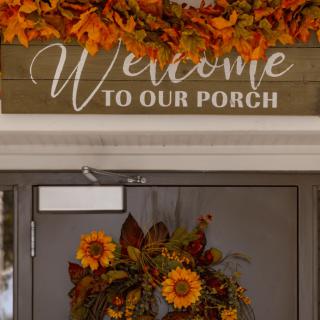 A cozy autumn-themed porch with a 'Welcome to Our Porch' sign and a sunflower wreath.