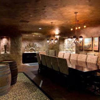 A luxurious wine cellar featuring a long dining table, surrounded by wine barrels and illuminated by elegant chandeliers.