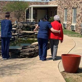 A healthcare worker supports an elderly person while walking outdoors near a building.
