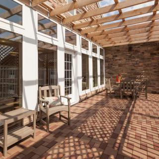 A sunlit patio with wooden furniture and a pergola overhead, featuring a brick floor.