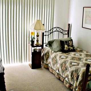 A cozy bedroom with a single bed, adorned with a patterned quilt, next to a bedside table and lamp. Vertical blinds cover the window area.