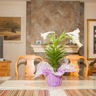 A dining room with a lily plant centerpiece on the table, surrounded by wooden chairs and decorated with artwork.