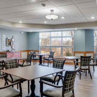 A well-lit dining room with tables and chairs arranged neatly, featuring large windows and a wooden floor.