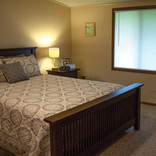 A cozy bedroom with a neatly made bed, bedside lamp, and a window letting in natural light.