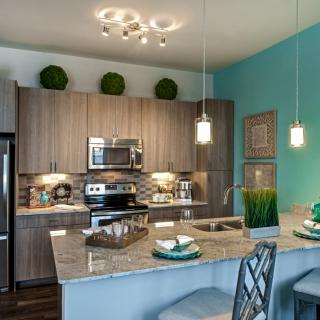 A modern kitchen featuring wood cabinets, granite countertops, and stainless steel appliances, accented by pendant lighting and decorative plants.