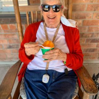 An elderly person wearing sunglasses and a red cardigan, enjoying a cup of ice cream while sitting on a rocking chair outdoors.