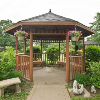 A wooden gazebo surrounded by lush greenery and flowers, with a stone bench nearby.