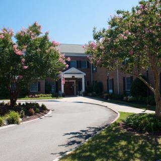 A large brick building with a circular driveway surrounded by well-maintained landscaping and trees on a sunny day.