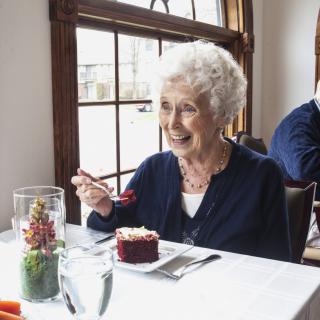 An elderly woman enjoying cake at a restaurant table by the window.