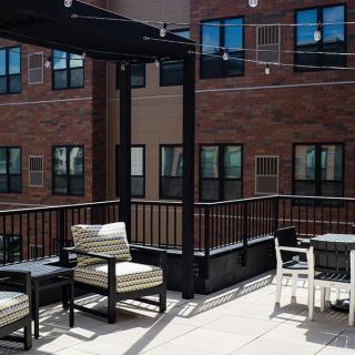 A cozy outdoor patio with seating, tables, and string lights against an apartment building backdrop.