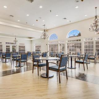 An elegant dining hall with rows of empty tables and chairs, illuminated by chandeliers and natural light from large windows.