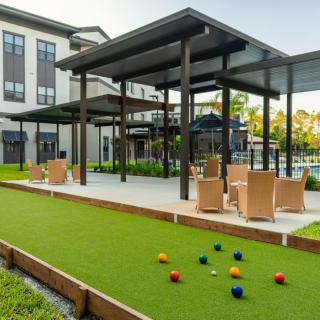 A modern outdoor patio with a pergola, seating area, bocce court, and landscaped surroundings next to a building.