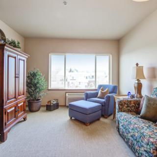 A cozy living room with a sofa, armchair, and armoire, featuring large windows and indoor plants.