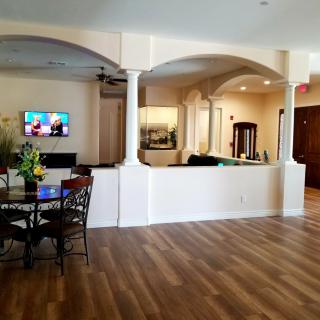 A spacious interior with a wooden floor, furnished with a table and chairs, featuring a wall-mounted television and decorative columns.