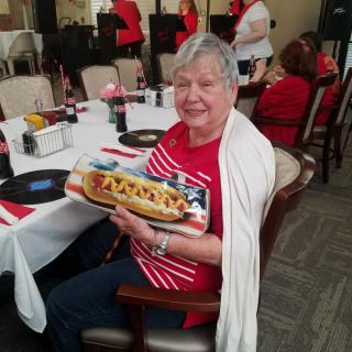An elderly woman smiling while holding a decorated hot dog at a party.
