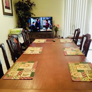 A dining room with a large wooden table, set with placemats and surrounded by chairs, facing a television.