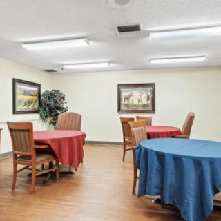 A well-lit dining room with tables covered in red and blue tablecloths, surrounded by wooden chairs and decorated with framed artwork on the walls.