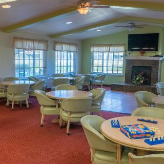 A spacious community room with tables and chairs, featuring a fireplace and large windows.