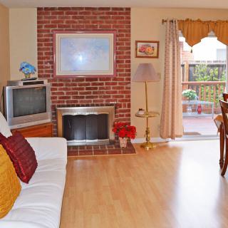A cozy living room featuring a brick fireplace, television, and a comfortable sofa, with an adjoining dining area leading to a patio.