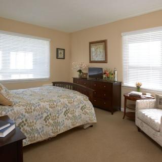 A cozy bedroom with a floral-patterned bedspread, a wooden dresser, and a comfortable chair by the window.