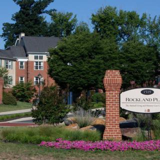 A scenic view of a brick building with a sign and well-maintained landscaping, featuring trees and colorful flowers.