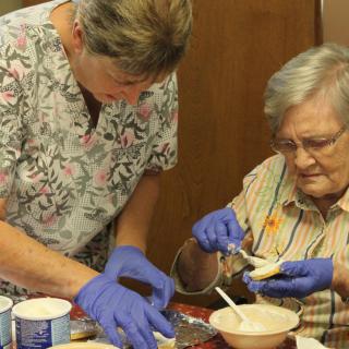 A caregiver assists an elderly woman with food preparation, both wearing gloves for hygiene.