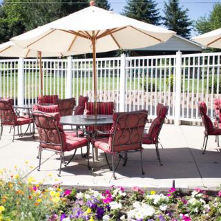 A sunny patio with tables and chairs under umbrellas, surrounded by colorful flowers and a white fence.