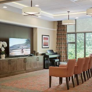 A well-decorated living room featuring a large TV, comfortable chairs, and a piano, with natural light streaming through large windows.