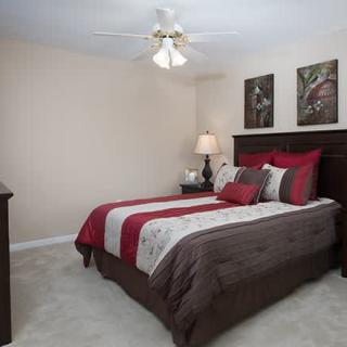 A cozy bedroom with a neatly made bed, featuring red and brown bedding, soft lighting from a table lamp, and decorative wall art.