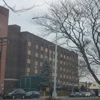 A view of a multi-story building surrounded by trees and parked cars, under a cloudy sky.