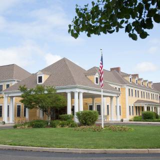 A large yellow building with a classical design, featuring columns and a flagpole with an American flag, set against a backdrop of trees and a partly cloudy sky.