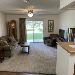 A cozy living room with a sofa, armchair, coffee table, and television, featuring a sliding glass door leading to a garden view.
