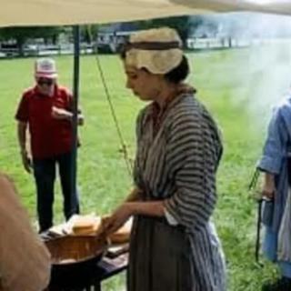 A group of people participating in a historical reenactment, with one person cooking outdoors in period costume.