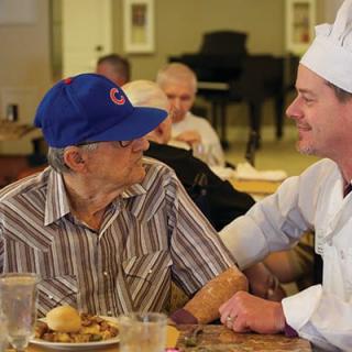 A chef engaging in a friendly conversation with an elderly man at a dining table.