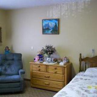 A cozy bedroom featuring a blue armchair, wooden dresser with decorative items, and a bed with floral bedding.