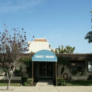 A small building with a blue awning labeled 'SUNSET MANOR', surrounded by trees on a clear day.