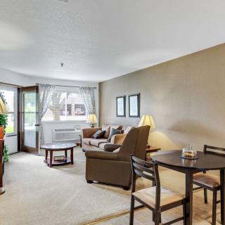A cozy living room with a sofa, table, chairs, and lamps, featuring warm lighting and a large window.