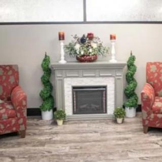 A cozy fireplace setup with two patterned armchairs, decorative plants, and candles in a well-lit room.