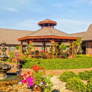 A beautifully landscaped garden featuring a central wooden gazebo surrounded by vibrant flowers and a multi-tiered fountain.
