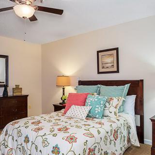 A cozy bedroom featuring a bed with colorful pillows and floral bedding, accompanied by a wooden dresser and a lamp, all under a ceiling fan.