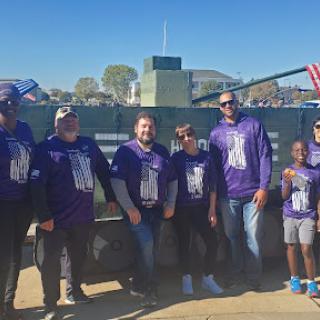 A group of friends and family gather for an outdoor event, all wearing matching purple shirts and enjoying the day.