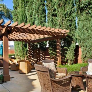 A sunny outdoor patio area with a wooden pergola and comfortable seating surrounded by lush greenery.