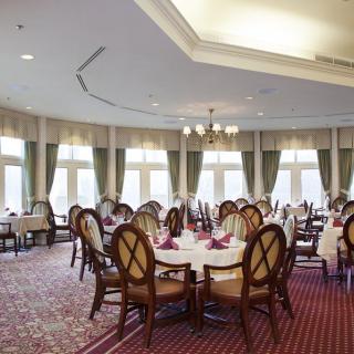 Elegant dining room with round tables and upholstered chairs, accented by large windows and a central chandelier.