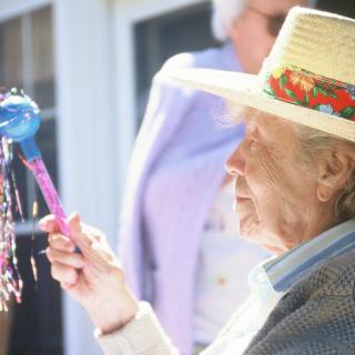 An elderly person wearing a hat is holding a colorful toy outdoors in the sunlight.