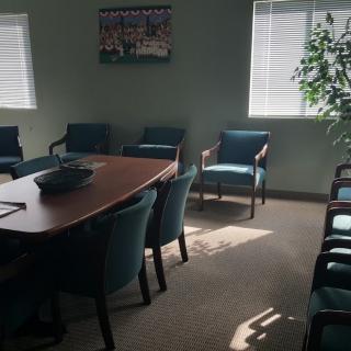 A conference room with a large table surrounded by chairs, a plant in the corner, and windows providing natural light.