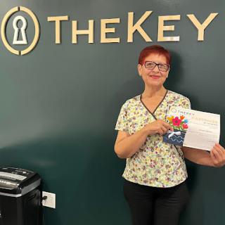 A smiling individual holding a document in front of a 'TheKey' sign in an office setting.